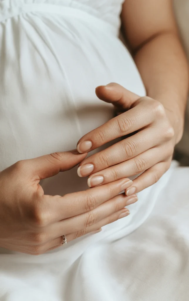 Nail care during pregnancy. Close-up of a woman's hands with smooth nails resting on her lap, wearing a white dress, elegant and serene.