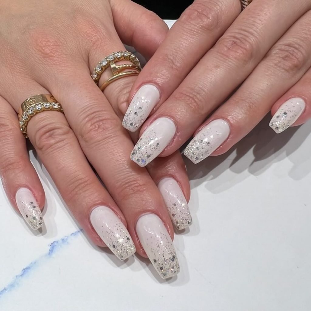 Elegant white nails with silver glitter and gold rings on hands resting on a white surface.