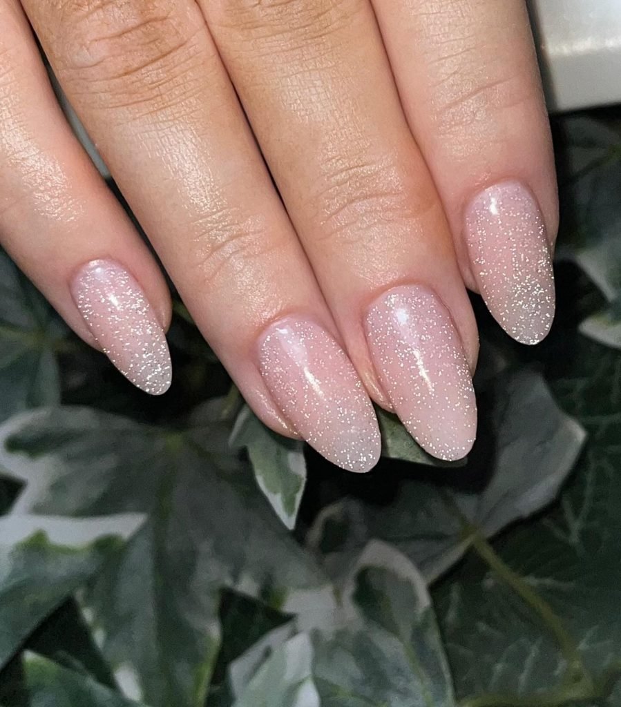 Glittery almond-shaped nails with soft pink polish and subtle sparkle, over green leaves background.