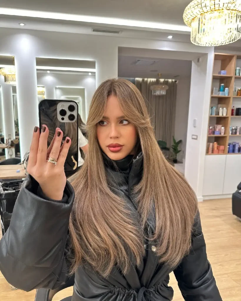 Young woman with long, straight hair in a salon mirror, wearing a black jacket and holding a phone for a selfie.