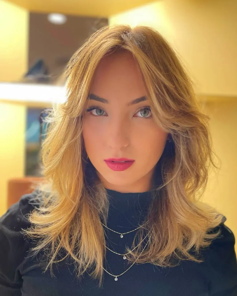 Portrait of a woman with long blonde hair and layered necklaces, featuring soft makeup and a vibrant background.