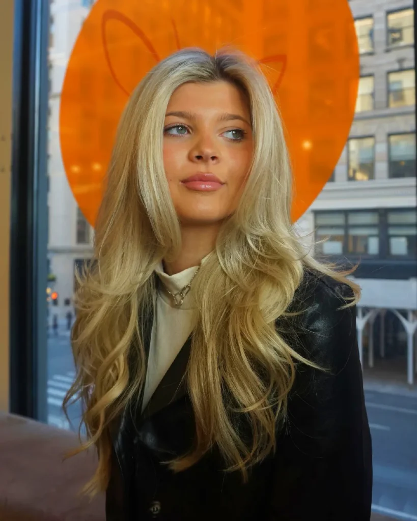 Blonde woman with long hair posing indoors near a large orange backdrop, wearing a black blazer and white top.