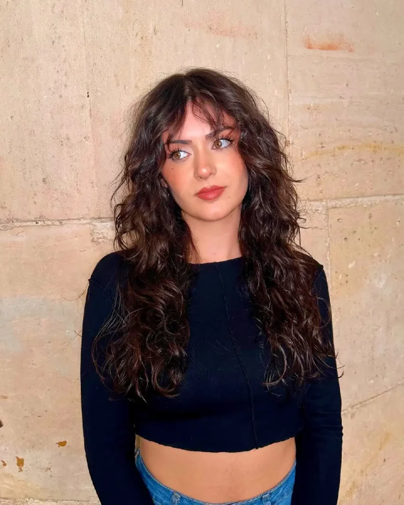 Young woman with long, curly hair wearing a black cropped top and blue jeans, posing against a textured wall.
