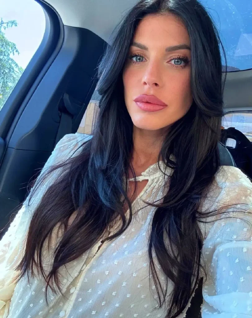 Close-up selfie of a woman with long dark hair, wearing a white blouse, sitting in a car with natural light.