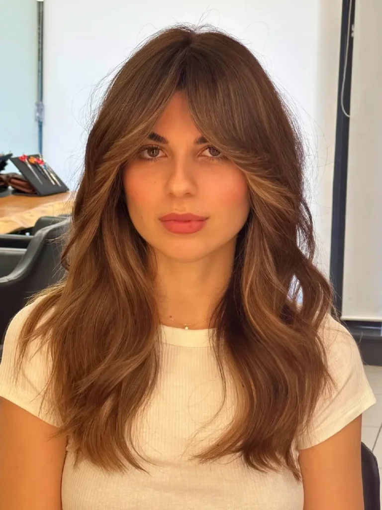 Woman with long, wavy brown hair and soft makeup, posing indoors in a bright, modern space.
