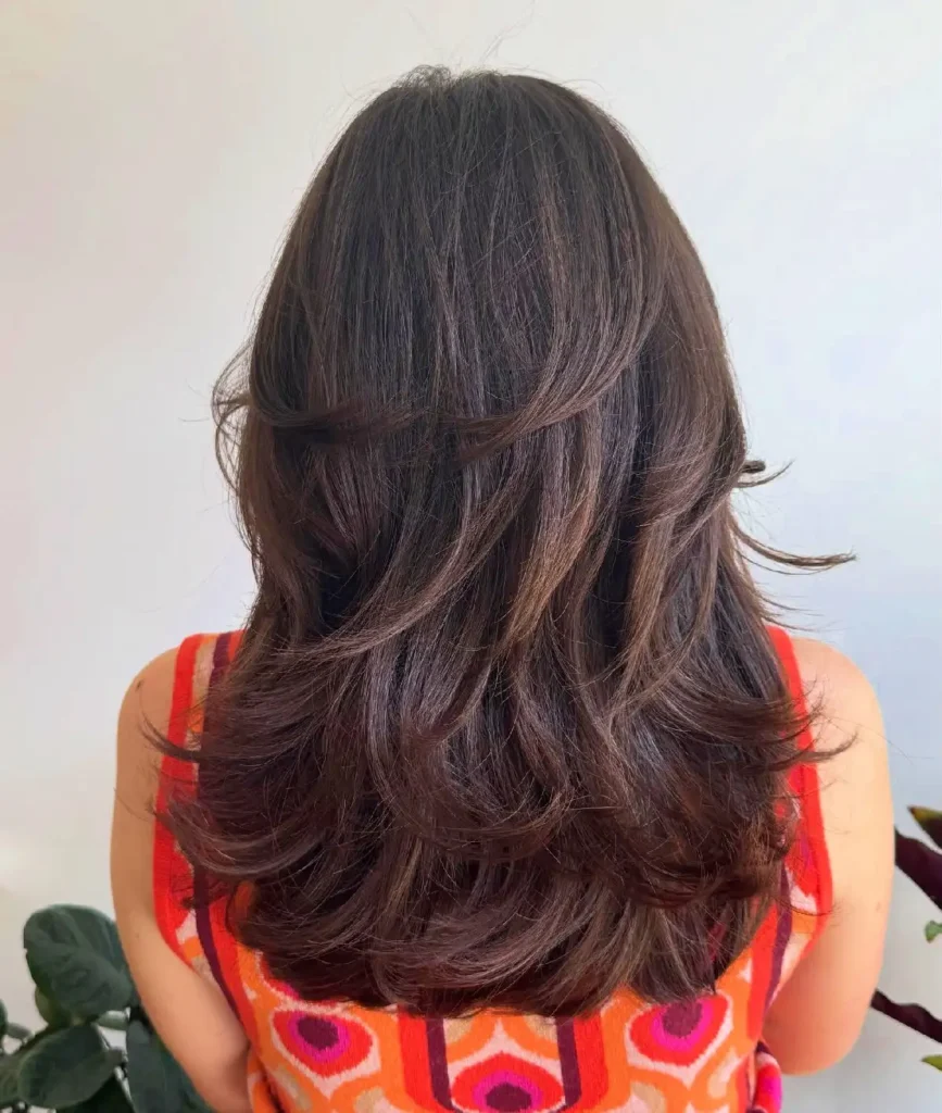 Woman with long, wavy brown hair, viewed from the back, wearing a colorful patterned top against a light background.