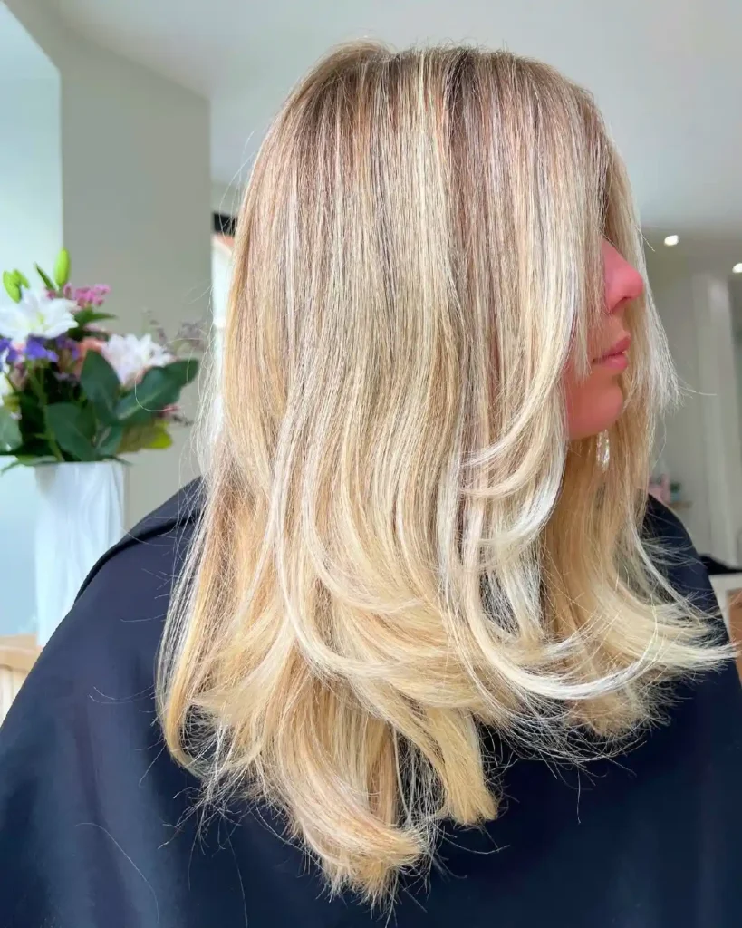 Fresh layered blonde hairstyle with soft waves, showcased in a bright salon setting with flowers in the background.