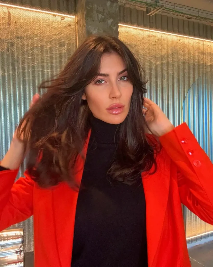 Young woman in a stylish red blazer and black turtleneck, posing with glossy hair in an urban setting.