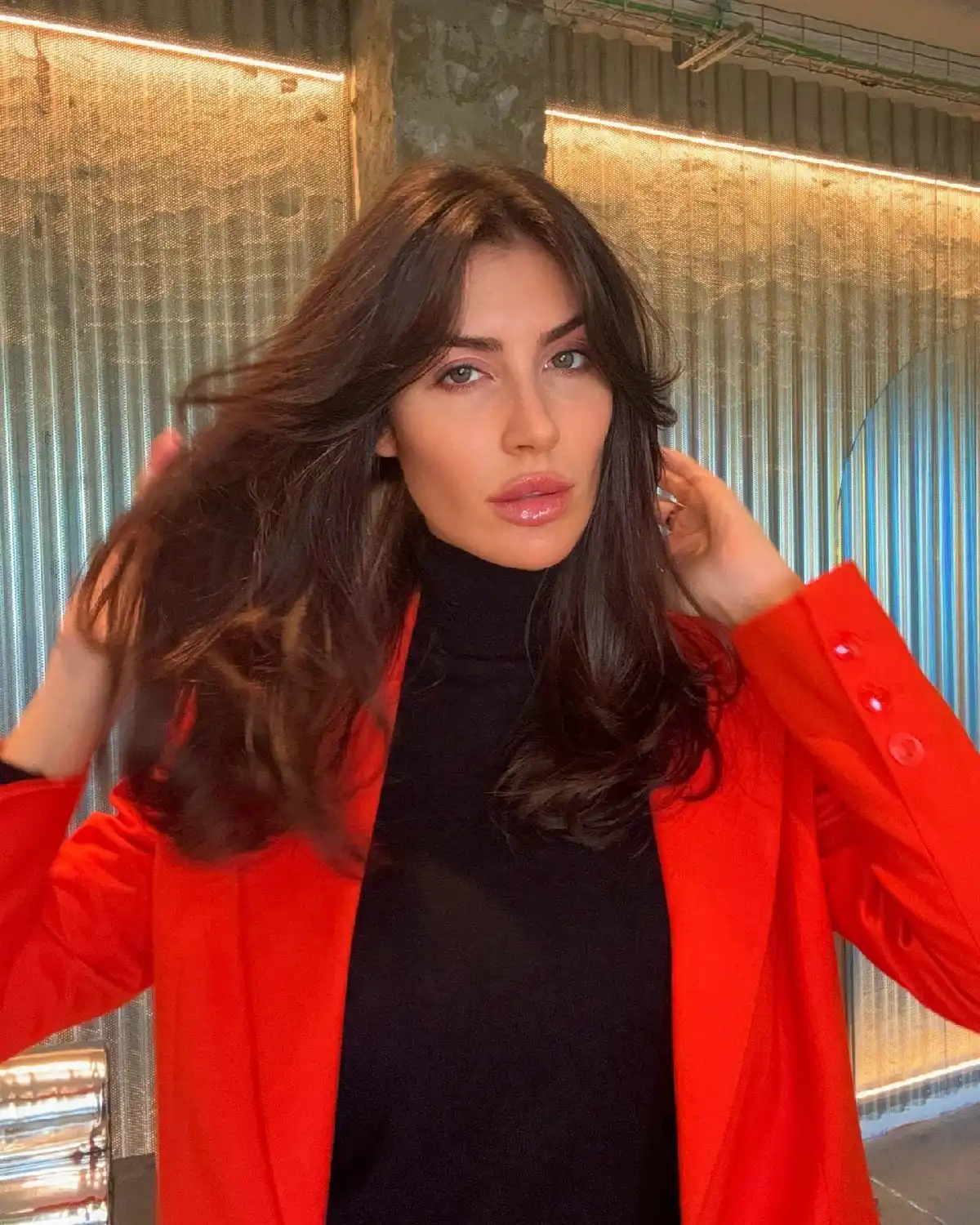 Young woman in a stylish red blazer and black turtleneck, posing with glossy hair in an urban setting.